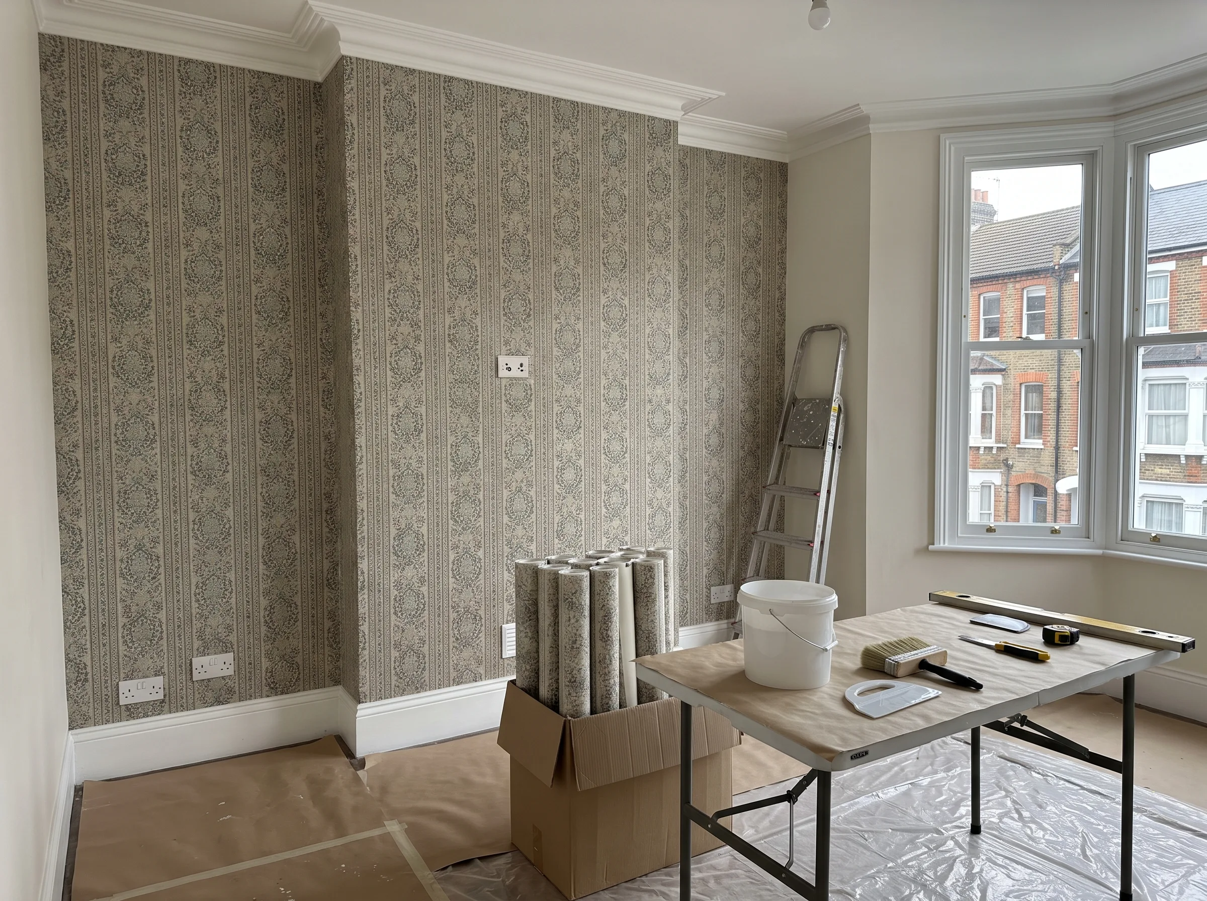 A room undergoing wallpapering with patterned wallpaper, a ladder, and tools on a table